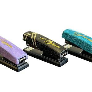 May include: Three staplers with glittered tops in purple, black, and teal. Each stapler has a name in gold script: Jessica, Alyssa, and Serena. The staplers are on a black base.