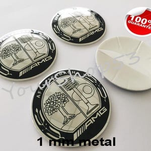 Puede incluir: Cuatro emblemas de coche de metal negro y plateado con el logotipo de Mercedes-Benz AMG y un diseño estilizado de árbol y pistón de motor. El texto "1 mm metal" está impreso debajo de los emblemas.