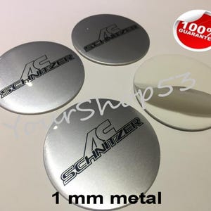 Pode incluir: Quatro tampas centrais de roda de carro de metal prateado com o texto preto "AC Schnitzer" em cada uma. As tampas têm 1 mm de espessura.