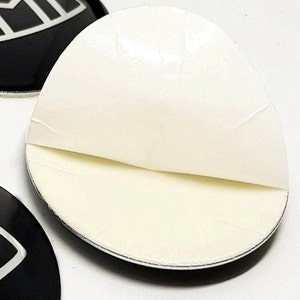 May include: A white adhesive backing is peeled back to reveal the sticky side of a round black sticker with a white design. The sticker is for decorative purposes.