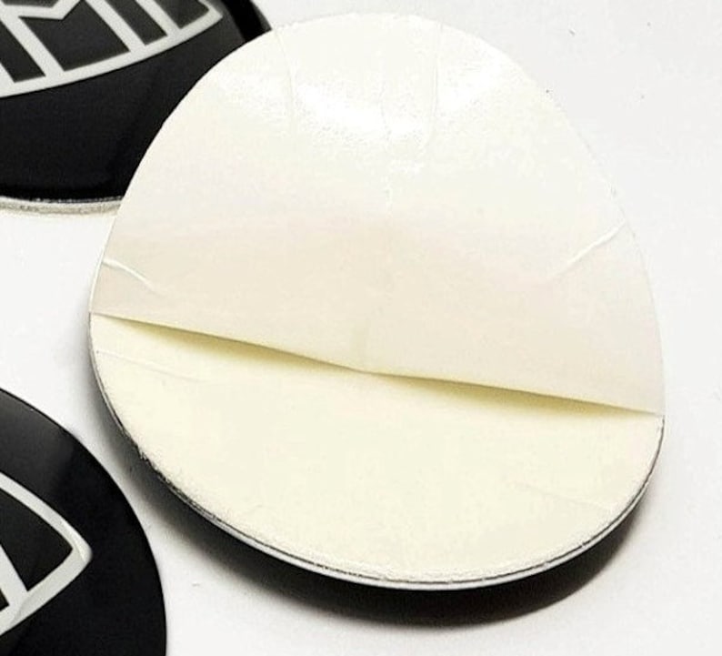 May include: A white adhesive backing is peeled back to reveal the sticky side of a round black sticker with a white design. The sticker is for decorative purposes.