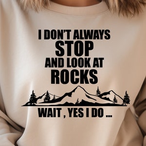 May include: A beige sweatshirt with a black graphic that reads "I don't always stop and look at rocks. Wait, yes I do..." with a mountain range and trees below the text.
