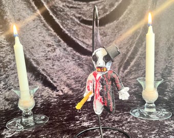 May include: A handmade Halloween decoration featuring a clown-like figure with a top hat, red and black suit, and a bloody face. The figure is mounted on a black metal stand, with two lit candles on either side.