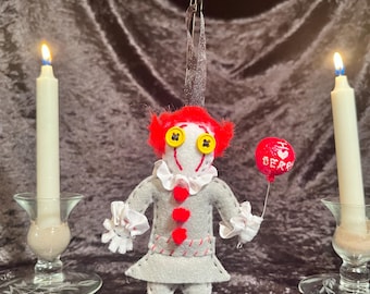 May include: A felt doll ornament of a clown with red hair, a white collar, and a gray dress. The doll is holding a red balloon with the text "I ♥ BERRY" on it. The doll is hanging from a metal stand against a dark brown background.