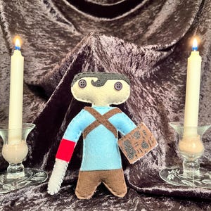 May include: A felt doll of a character wearing a blue shirt with a red and white arm, brown trousers, and a black and white mask. The doll is holding a book with black and white text. The doll is sitting on a brown velvet surface with two lit candles on either side.