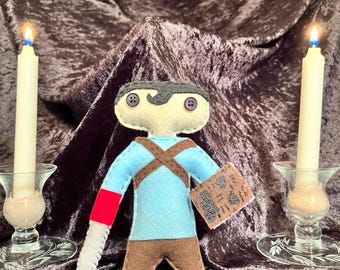 May include: A felt doll of a character wearing a blue shirt with a red and white arm, brown pants, and a black and white mask. The doll is holding a book with black and white text. The doll is sitting on a brown velvet surface with two lit candles on either side.