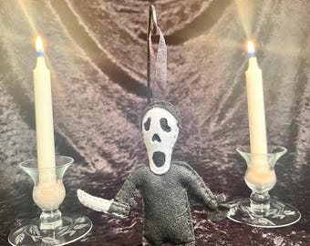 May include: A felt Scream mask ornament with a black body and white face, arms outstretched. It is held by a metal clip. Two lit candles in glass holders flank the ornament, set against a dark, textured background.