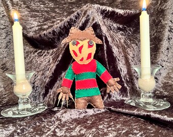 May include: A felt doll of the iconic horror movie character Freddy Krueger. The doll is wearing a brown hat, a red and green striped sweater, and brown pants. The doll has a red and orange mask with a scary face. The doll is sitting on a brown velvet surface with two lit candles on either side.