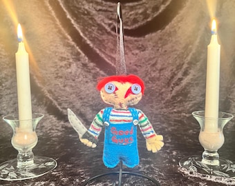 May include: A handmade felt doll ornament of a red-haired boy with a blue shirt and overalls. The doll is holding a knife and has the words "Good Guys" embroidered on the front. The doll is on a black metal stand and there are two lit candles in the background.