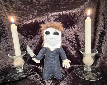 May include: A felt doll of a horror movie character wearing a white mask and holding a knife. The doll is standing on a dark velvet surface with two lit candles on either side.