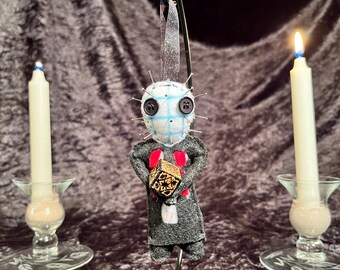 May include: A handmade ornament featuring a character with a blue and white checkered face, black button eyes, and pins. The character wears a gray coat with a red heart and holds a small, patterned cube. Two lit candles flank the ornament.