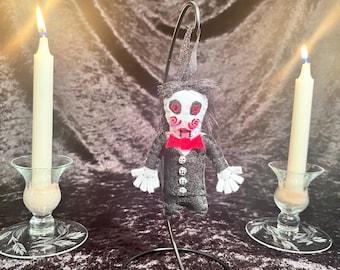 May include: A handmade felt doll ornament of a horror movie character with a black suit, white gloves, and a red bow tie. The doll is hanging from a black metal stand. Two white candles are in the background.