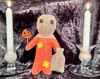 May include: A handmade burlap doll with a red and orange outfit, holding a stick with a pumpkin-shaped head. The doll is standing on a black velvet surface with two white candles in glass candle holders on either side.