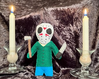 May include: A felt doll of a horror movie character wearing a white mask with red accents and a green shirt, holding a knife and an axe. The doll is standing on a dark brown velvet surface with two lit candles on either side.