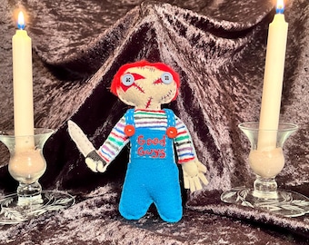 May include: A handmade felt doll with red hair, stitched-on eyes, and a knife. The doll is wearing a blue and white striped shirt, blue overalls, and has the words "Good Guys" embroidered on the front. The doll is sitting on a dark brown velvet surface with two lit candles on either side.