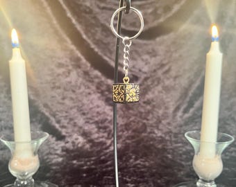 May include: A black and gold cube keychain with intricate details hangs from a silver chain on a black metal stand. Two white candles are in the background.
