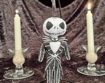 May include: A felt Jack Skellington ornament, featuring a white head with black eye and mouth details, and a black and white pinstripe suit. The ornament hangs from a metal loop. Two lit candles in glass holders flank the ornament.