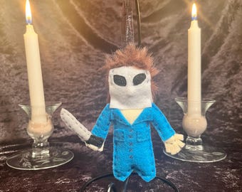 May include: A handmade felt ornament depicting a character in a blue jumpsuit, holding a knife. The ornament has a white mask and brown hair. Two lit white candles in glass holders flank the ornament, creating a spooky Halloween theme.