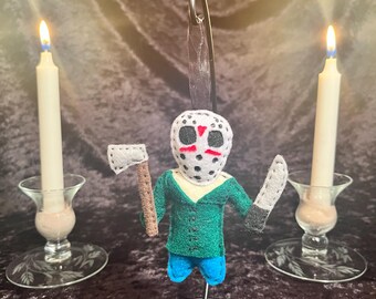 May include: A felt ornament of a horror movie character wearing a white mask with red and black details, holding a knife and an axe. The ornament is hanging from a black metal stand.