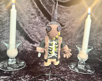 May include: A brown felt doll with a skeleton costume and yellow button eyes. The doll is hanging from a black metal stand against a dark brown velvet background.
