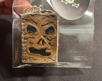 May include: A tan, textured keychain shaped like a book with a face, featuring black recessed eyes and a mouth, attached to a silver metal ring. A circular sticker is included in the clear plastic packaging with a skull and the text Julie's Sinister Shop.