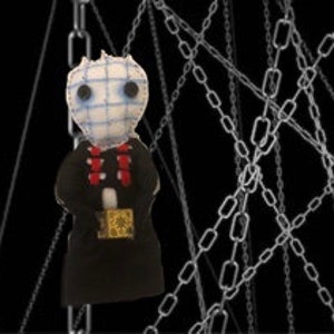 May include: A handmade doll with a white fabric face and black eyes, wearing a black dress and a red necklace. The doll is holding a small book and is surrounded by silver chains.