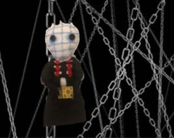 May include: A handmade doll with a white fabric face and black eyes, wearing a black dress and a red necklace. The doll is holding a small book and is surrounded by silver chains.