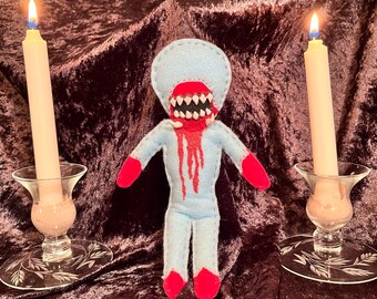 May include: A blue felt doll with a red mouth and dripping red paint. The doll is standing between two lit candles in glass candle holders on a brown velvet surface.