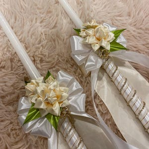 May include: Two white candles decorated with ivory satin ribbons, bows, and floral accents. The candle handles are wrapped with ivory satin and embellished with rhinestones. Green leaves and gold accents complete the design.