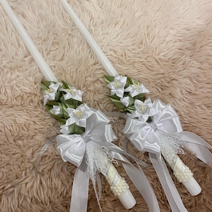 May include: Two white taper candles decorated with white satin bows, lace, and faux pearls. The candles are adorned with white fabric flowers and green leaves. The candles are for a wedding or special occasion.
