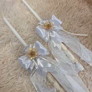 May include: Two white candles decorated with silver ribbons, lace, and a gold flower embellishment. The candles have a satin bow and long, flowing ribbons. The candles are likely for a special occasion.