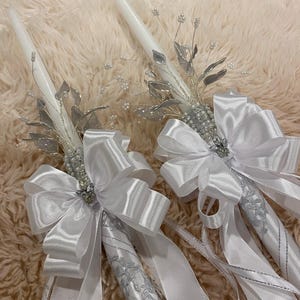 May include: Two white taper candles decorated with silver leaves, beaded accents, and large white satin bows. The candles have long white ribbons and are designed for a special occasion.