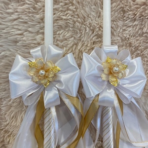 May include: Two white candles decorated with white satin bows, gold ribbons, and floral accents. The candles have gold and white striped patterns and are adorned with faux pearls. A decorative item for special occasions.