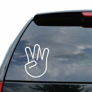 May include: White vinyl decal of a hand making the 'W' sign, commonly used to represent the number three. The decal is on the back window of a black SUV.