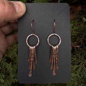 May include: Copper-colored dangle earrings. Each earring features a hammered copper ring with multiple hammered copper fringe elements hanging below. The earrings are displayed on a black card against a backdrop of green foliage.
