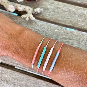 May include: Five colorful beaded bracelets on a person's wrist. The bracelets are made with small, round beads in shades of pink, white, and turquoise. The bracelets are tied with thin, pink, white, and turquoise cords.