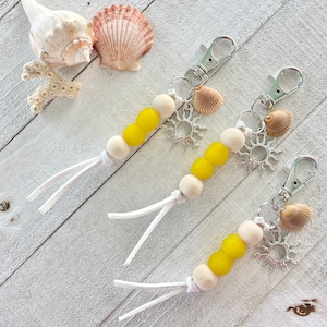 May include: Three keychains with yellow and white beads, silver charms, and seashell accents. The charms include a sun and a seashell.