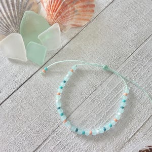 May include: A delicate beaded bracelet with light blue, peach, and dark blue beads. The bracelet is on a light green string. Seashells and sea glass are in the background on a white wooden surface.