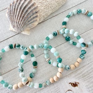 May include: Four beaded bracelets with a beachy theme. The bracelets feature a mix of turquoise, white, and wooden beads. A seashell and a piece of coral are also present, all arranged on a white wooden surface.