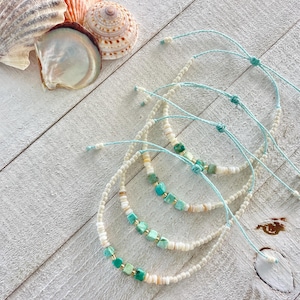May include: Several handmade anklets with turquoise and white beads, and adjustable light blue cords. The anklets are adorned with small gold-colored beads and square turquoise stones. Seashells are scattered on a white wooden surface.