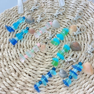 May include: Several keychains with silver clasps, featuring sea glass beads in shades of blue, green, and pink. Each keychain is adorned with a small seashell. The keychains are arranged on a woven straw background.