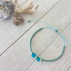 May include: A bracelet featuring a light blue cord with small sage green seed beads and two larger, smooth, bright blue glass beads. The bracelet has an adjustable sliding knot closure with two loose cord ends extending from the knot.