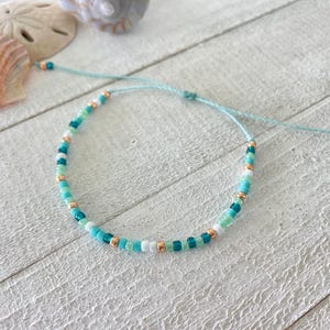May include: A delicate beaded bracelet with a mix of turquoise, white, and gold beads. The bracelet is tied with a thin blue cord.