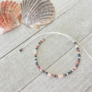 May include: A delicate bracelet featuring small, multi-colored glass beads in shades of cream, dark blue, and terracotta, strung on a thin white cord with adjustable ends, positioned near two large, textured seashells.