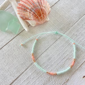 May include: A beaded bracelet with a light blue cord, featuring light blue, peach, and gold-colored beads. A starfish, seashell, and sea glass are in the background, all on a white wooden surface. The bracelet is adjustable.