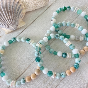 May include: Three beaded bracelets with a beach theme. The bracelets feature a mix of white, teal, and green beads, along with wooden and shell accents. A seashell is in the background.