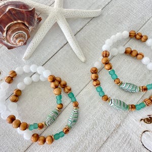 May include: Two beaded bracelets with white, brown, and teal beads. The bracelets are next to a seashell and a starfish on a white wooden surface. The bracelets have a beachy, coastal theme.