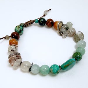May include: A beaded bracelet featuring a variety of natural stone beads in shades of brown, green, and clear. The bracelet includes a mix of round and oblong beads, with a macrame closure. The bracelet is on a white background.