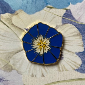 May include: A blue enamel pin with a white flower with a yellow center. The pin is shaped like a flower.