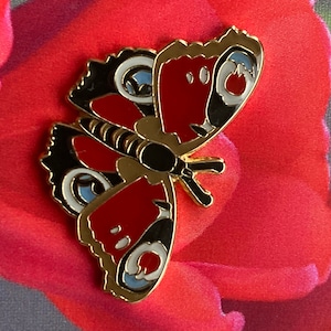 May include: A gold-colored metal pin with a red, black, and blue butterfly design. The butterfly has white spots on its wings.